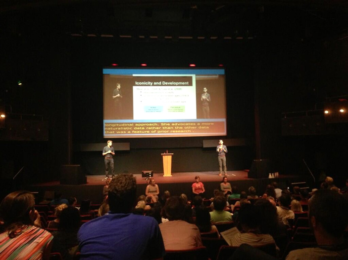 Выступление на конференции «Теоретические вопросы исследования жестовых языков». Докладчик (справа) говорит на амслене, его переводят на британский (на сцене слева) и другие жестовые языки (под сценой)