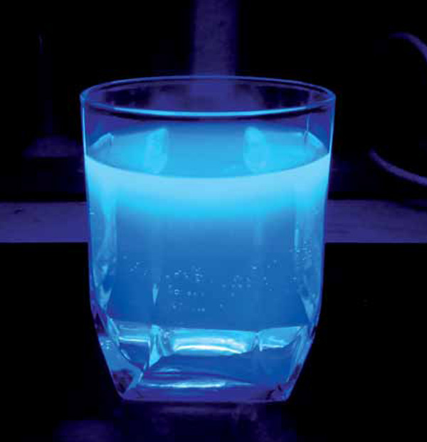 Рис. 1. Раствор сульфата хинина в стакане облучается сверху ультрафиолетом (фото автора). Эта фотография иллюстрирует целых два физических закона. Во-первых, УФ-излучение поглощается раствором хинина, и интенсивность окраски уменьшается сверху вниз — это следствие закона Бугера—Ламберта—Бера. Во-вторых, если облучать стакан не сверху, а сбоку, то флуоресценции не будет, так как стекло не пропускает ультрафиолет. При этом свет, испускаемый раствором хинина, через стекло проходит. Это означает, что длина волны излученного света больше длины волны поглощенного света — закон Стокса. Изображение: «Химия и жизнь»